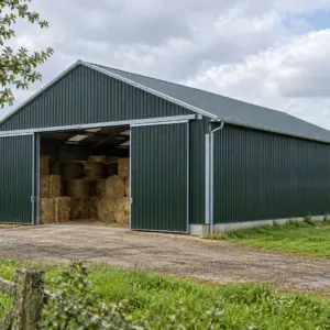Hangar agricole bardé de tôle verte, porte coulissante ouverte sur des balles de paille et de foin, en Normandie